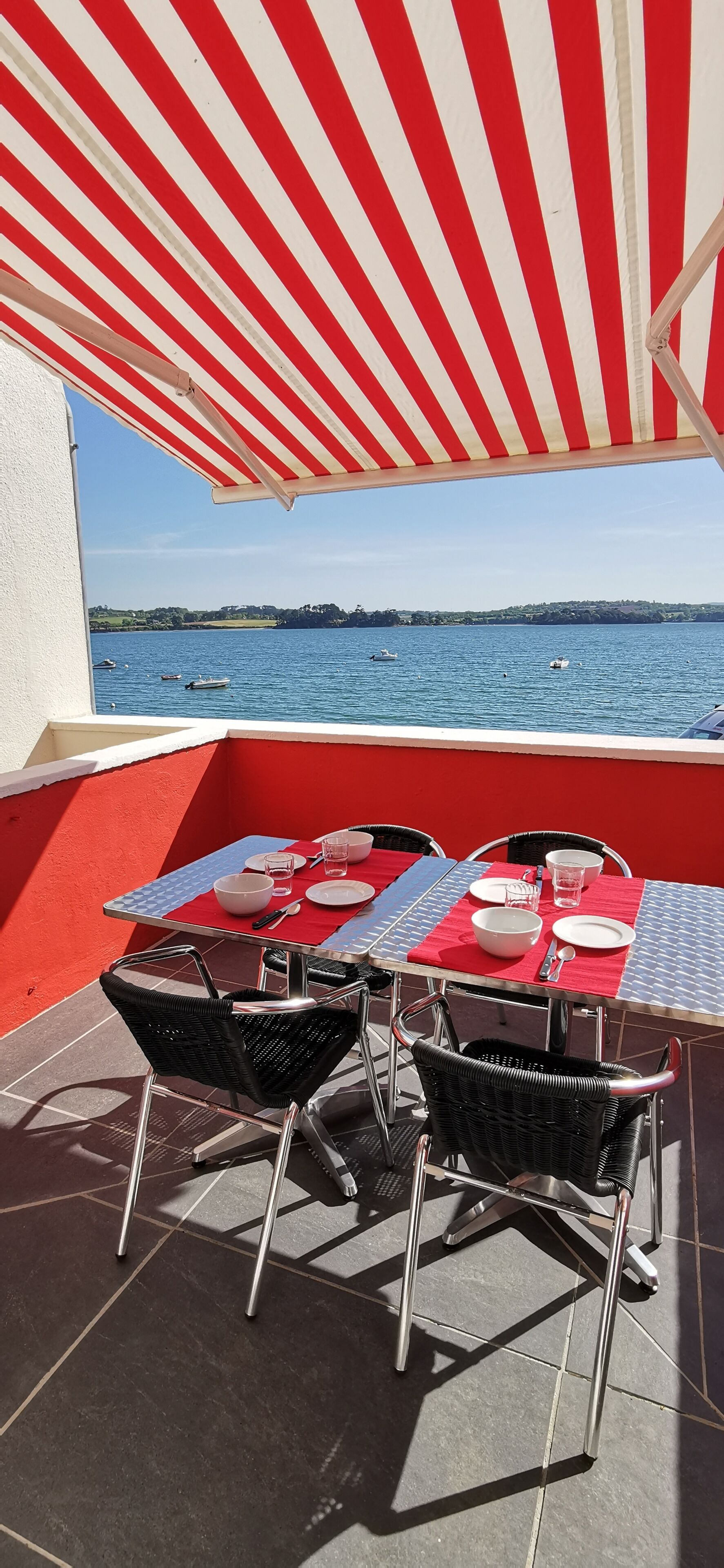 Un área de comedor al aire libre con vista al mar, con toldos a rayas rojas y blancas, mesas de metal y sillas de mimbre.