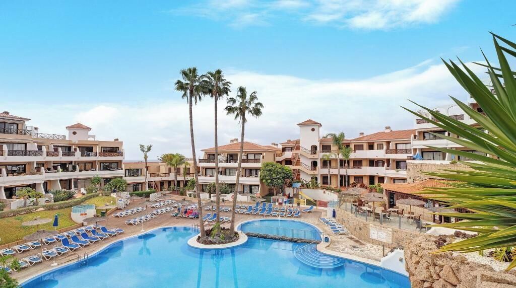 A hotel with a central swimming pool, multiple balconies, and palm trees.