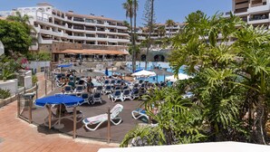 Poolanlage eines Resorts mit Liegestühlen und Sonnenschirmen, umgeben von Palmen und Hotelgebäuden.