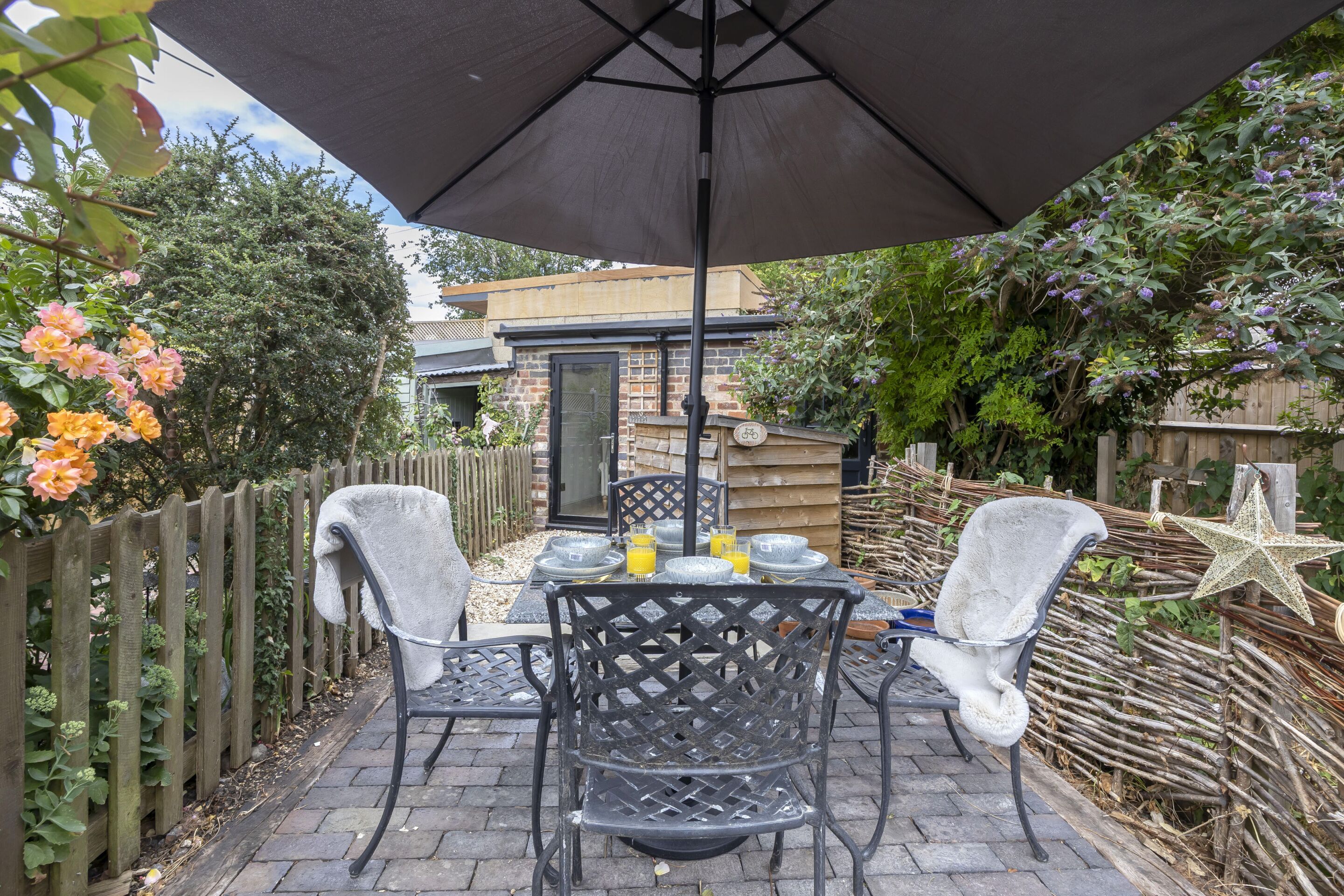 An outdoor patio with a table set for two, surrounded by chairs, under a large umbrella.