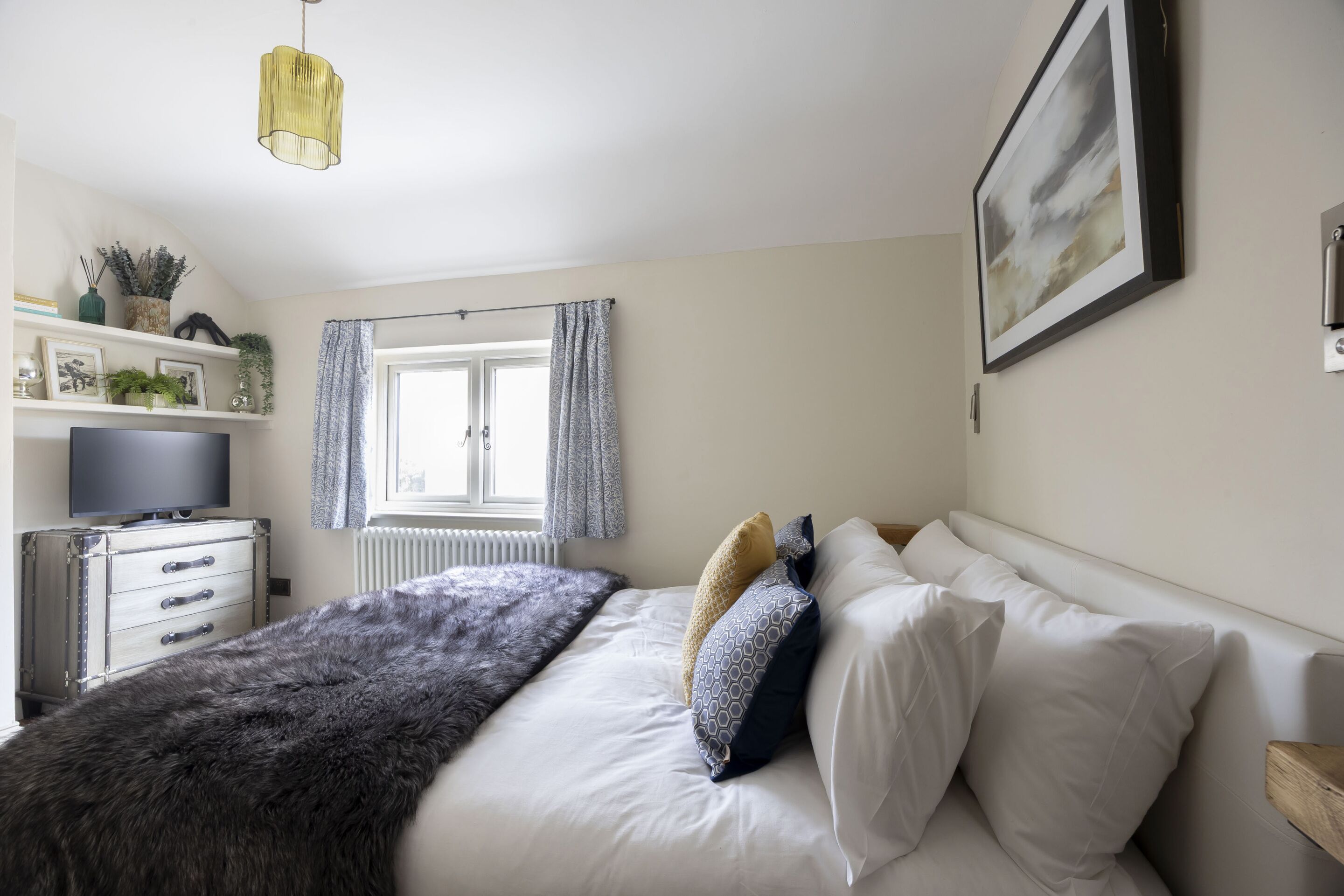 Een slaapkamer met een bed, een dressoir, een televisie, een radiator, een raam met gordijnen en een schilderij aan de muur.