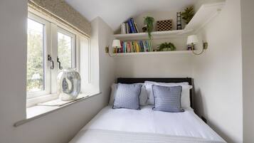 A bedroom with a bed, a window with a view, a shelf with books and plants, and a wall-mounted lamp.