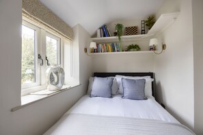 Una camera da letto con un letto, una finestra vista, uno scaffale con libri e piante, e una lampada a parete.