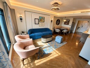 Ein modernes Wohnzimmer mit einem blauen Sofa, einem pinken Sessel und einem Couchtisch. Es gibt einen Essbereich mit Tisch und Stühlen, sowie eine Küche im Hintergrund.