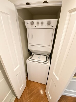 A compact, white Whirlpool washer and dryer unit installed in a narrow hallway with wooden flooring. - Orlando | Family Riviera Retreat | Pool + Resort | 2BR (Lake Buena Vista)