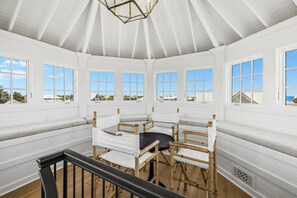 Ein Zimmer mit hohen Decken, weißen Wänden und großen Fenstern. In der Mitte steht ein Holztisch mit Stühlen und einem schwarzen Kissen.