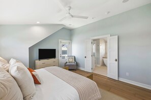 Ein Schlafzimmer mit Bett, Fernseher, Deckenventilator und einem durchgehenden Türrahmen sichtbaren Badezimmer.