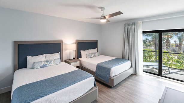 A hotel room with two beds, a ceiling fan, a nightstand with a lamp, and a view of the outdoors.