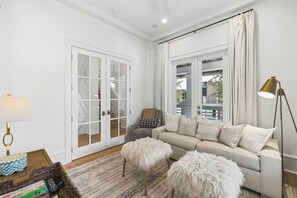 A living room with a sofa, two fluffy ottomans, a lamp, and a view through a glass door. - Porches | Gulf Views | Carriage House (Rosemary Beach)