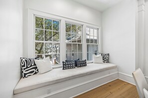 A room with a built-in bench along a white wall, large windows with multiple panes, and a view of trees outside. - Porches | Gulf Views | Carriage House (Rosemary Beach)