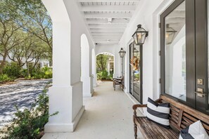 Un porche cubierto con un banco, una puerta adornada con un arreglo floral y vista a un jardín.