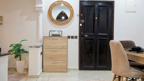 Una habitación con una puerta de madera oscura, un armario de madera con cajones, un espejo redondo en la pared, una planta en maceta y una silla color beige.