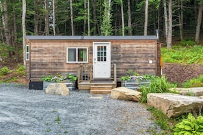 A small wooden cabin with a white door, two windows, and a gravel driveway.