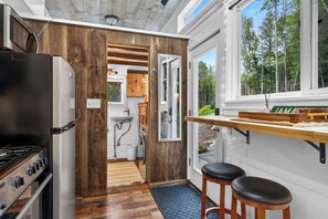 A compact kitchen with a wooden interior, a refrigerator, a sink, and a window overlooking a forest.