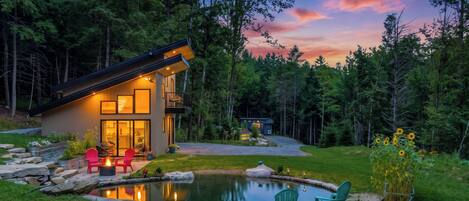 Une cabane moderne dotée d’une entrée en verre, d’un espace extérieur aménagé et d’un étang.