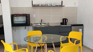 A compact kitchen with a built-in oven, a sink, and a kettle on the countertop. There is a round glass table with four yellow chairs.