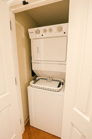 Een kleine wasruimte met een gestapelde wasmachine en droger, een klein schap met flessen, en een deur naar een andere ruimte.