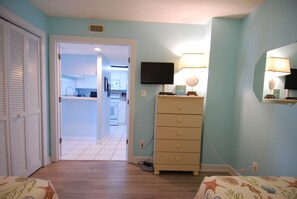 A room with a wooden floor, a dresser, a television mounted on the wall, and a door leading to a kitchen. - 3BR Oceanfront Condo at Land's End! (Myrtle Beach)