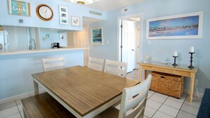 A dining area with a wooden table and chairs, a kitchen area with a coffee maker, and a wall with framed pictures and a clock. - 3BR Oceanfront Condo at Land's End! (Myrtle Beach)