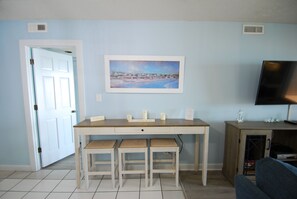 Een kamer met een houten eettafel, twee stoelen, een schilderij aan de muur, een televisie op een standaard, en een deur naar een andere kamer.