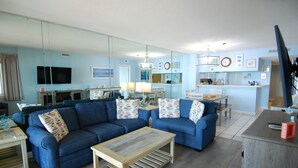 A modern living room with a blue sofa set, a wooden coffee table, and a kitchen area with a dining table. - 3BR Oceanfront Condo at Land's End! (Myrtle Beach)