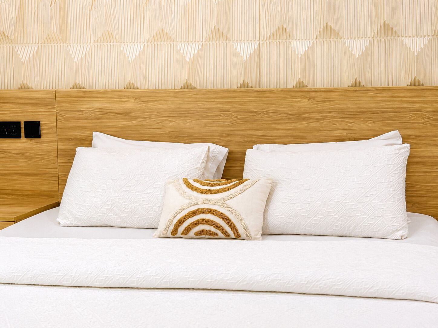 A neatly made bed with white bedding and decorative pillows, set against a wooden headboard and patterned wall.