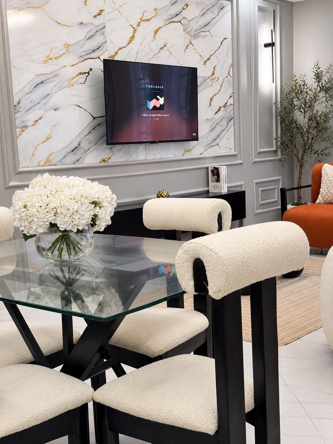 A modern dining area with a glass table, cushioned chairs, a marble wall, and a flat-screen TV displaying a music video.