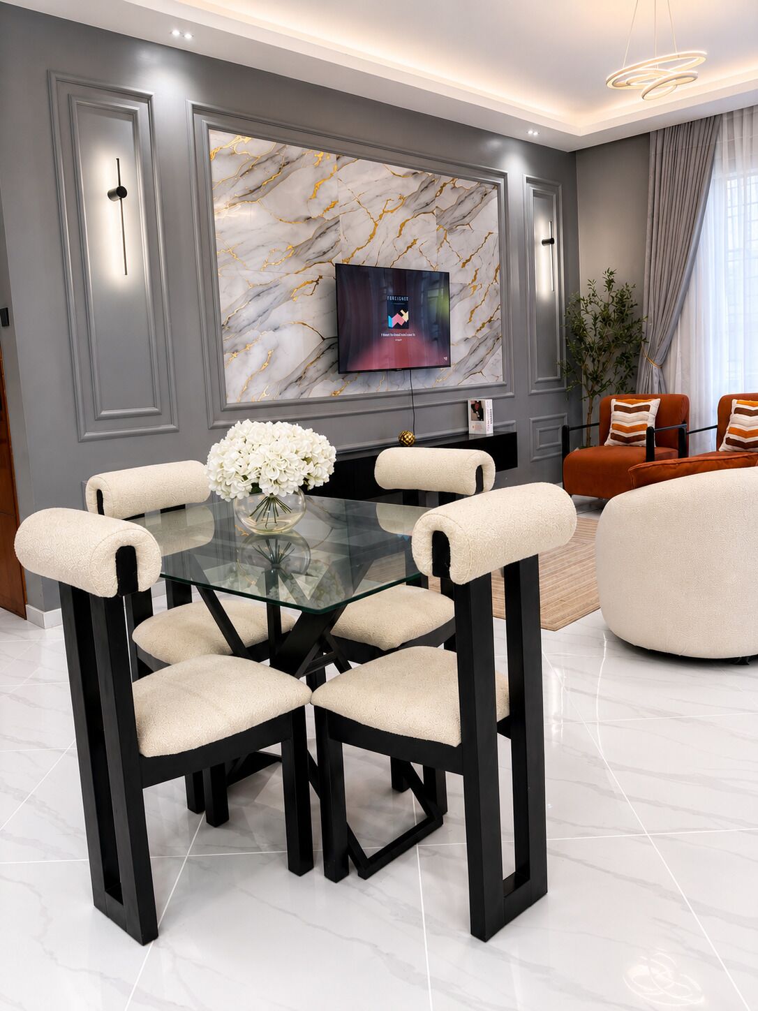 A modern dining area with a glass table and chairs, a marble wall with a TV, and a plant.