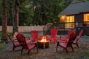 Ein Feuerkorb mit vier Adirondack-Stühlen, die darum platziert sind, auf einem Kiesgrundstück, Bäume im Hintergrund.