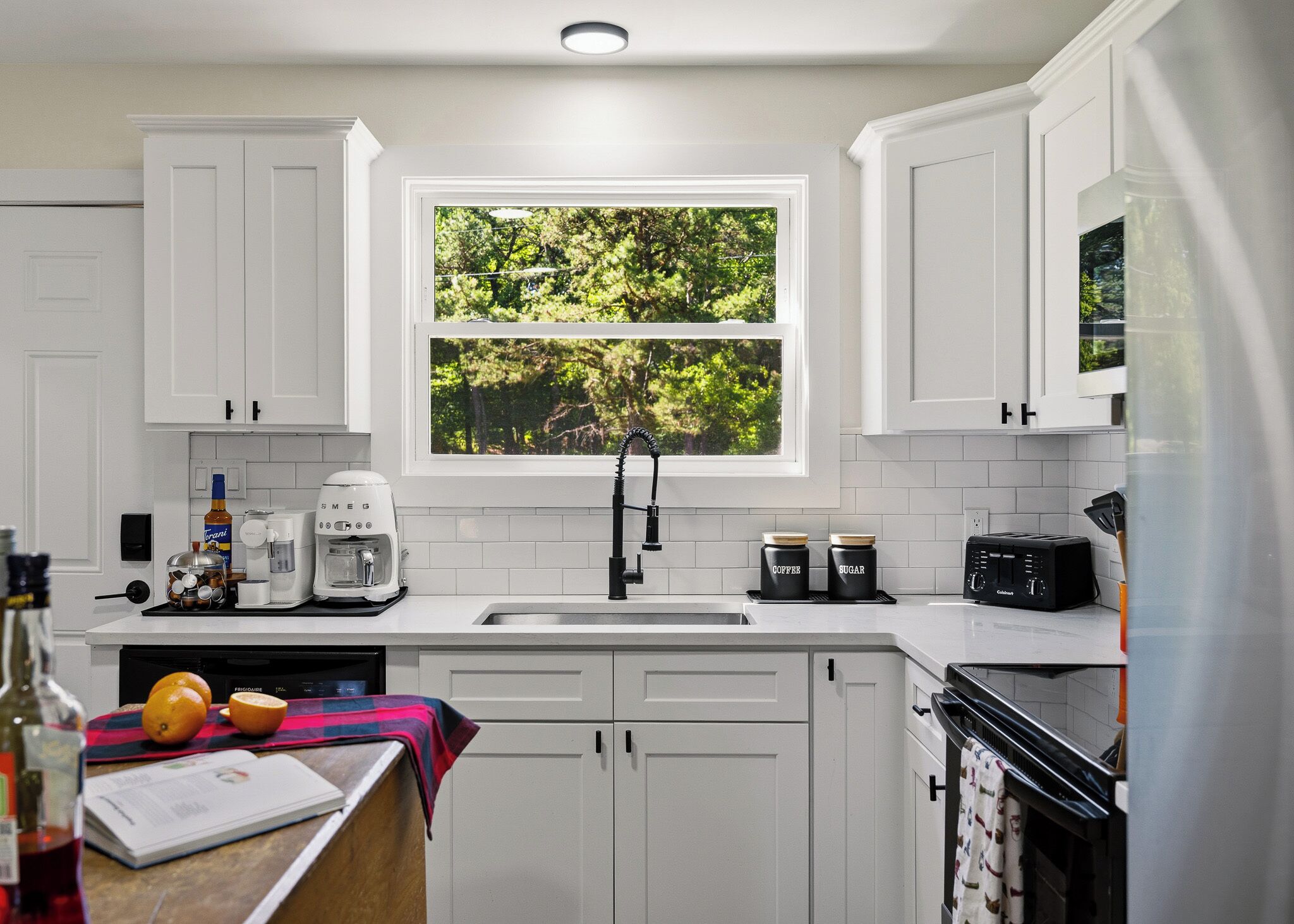 A modern kitchen with white cabinets, a window with a view of greenery, a coffee maker, a toaster, and a refrigerator.
