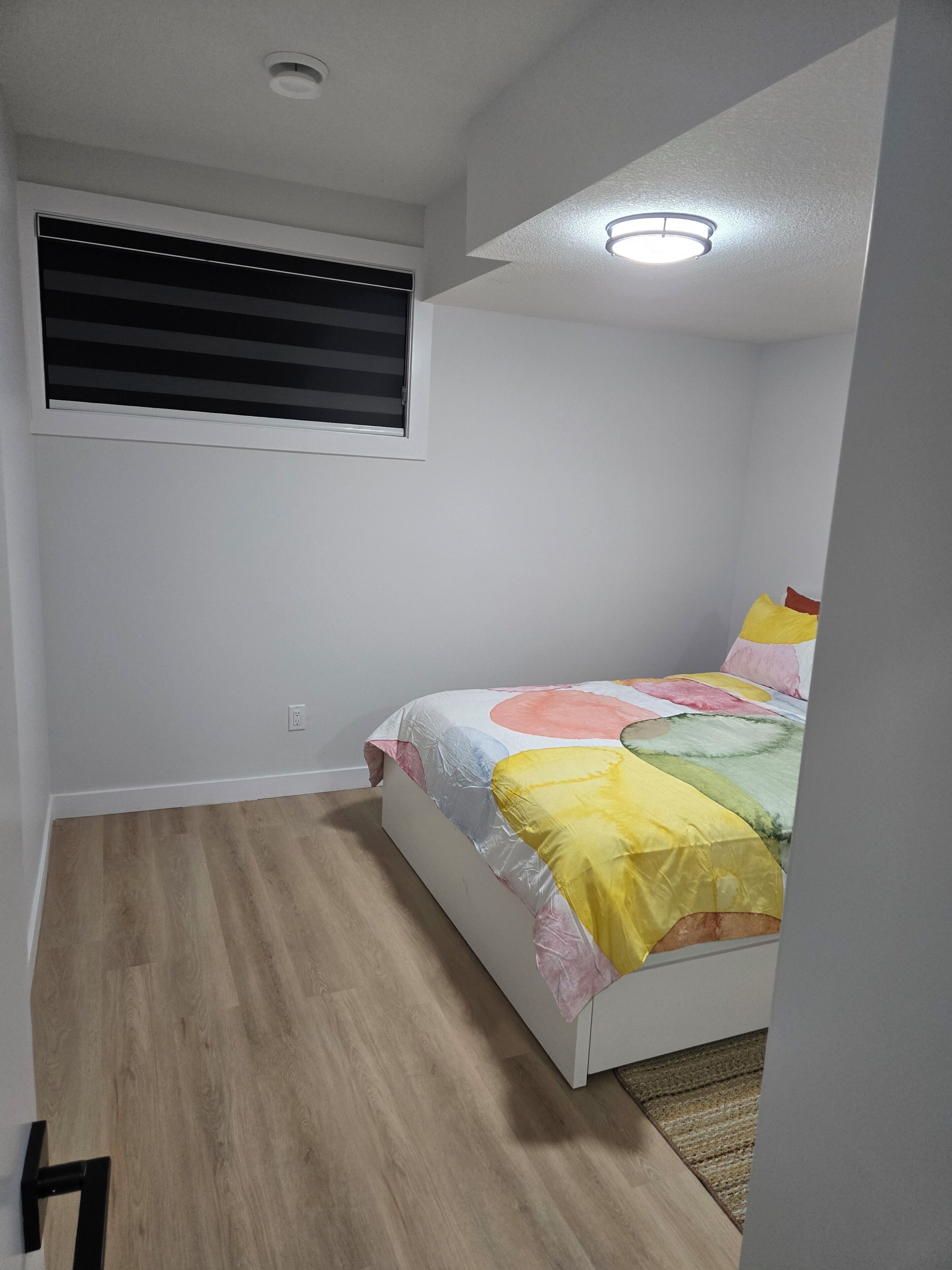 Un dormitorio con cama, ventana con persianas, luz de techo y suelo de madera.