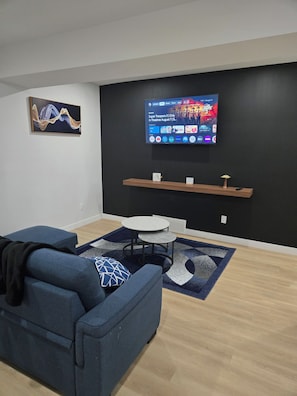 A modern living room with a blue sofa, a small round table, a flat-screen TV mounted on the wall, and a decorative wall piece. - Imperial Lodge:3BD Peaceful Getaway Suite+King Bed (Edmonton)