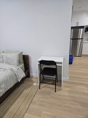 A bedroom with a bed, a desk, and a chair. There is a kitchen area with a refrigerator in the background. - Imperial Lodge:3BD Peaceful Getaway Suite+King Bed (Edmonton)