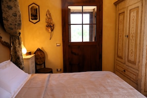 Una habitación con una cama, una puerta de madera, una silla, un escritorio con una lámpara y un cuadro colgado en la pared.