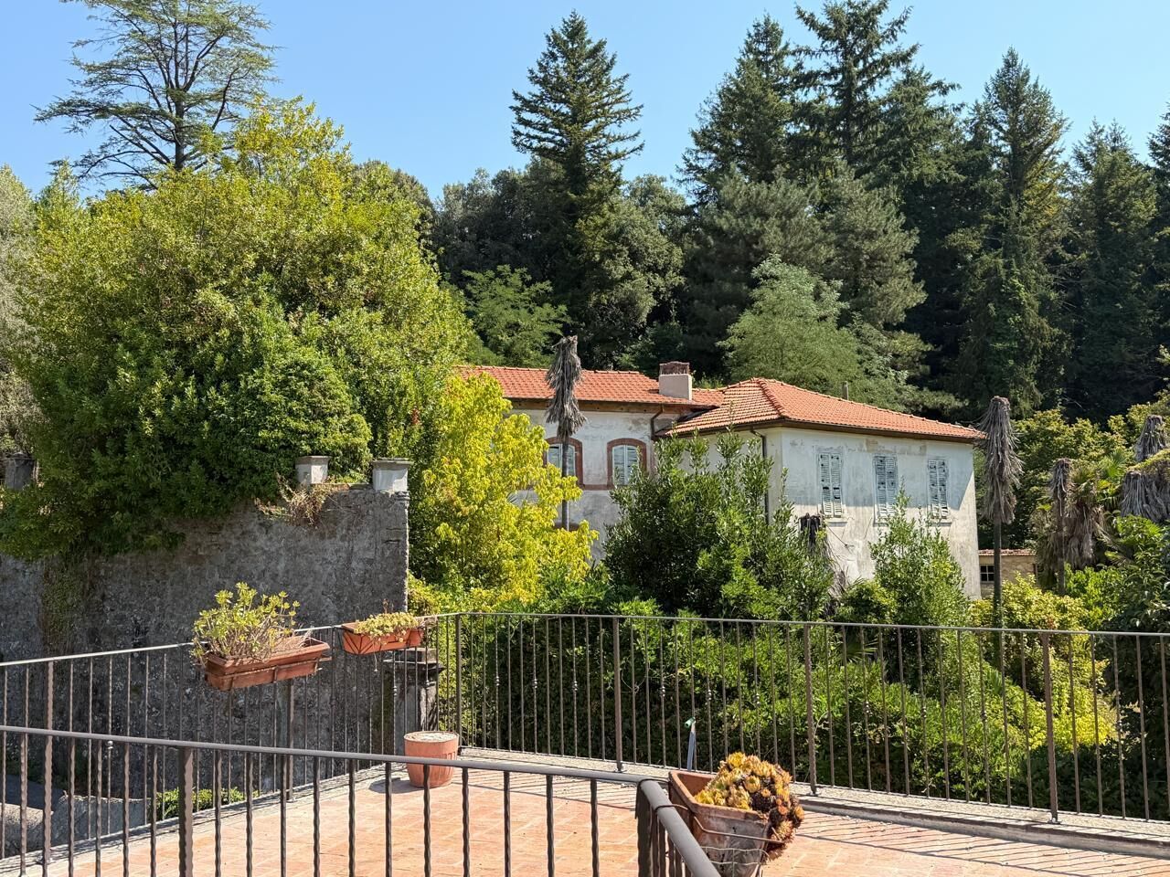 A balcony with potted plants, a stone wall, and a building with a red-tiled roof in the background.