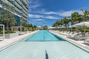 A swimming pool with lounge chairs and umbrellas, surrounded by palm trees and modern buildings.
