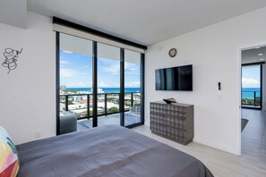A modern bedroom with a large bed, a TV mounted on the wall, and a view of the city and ocean through floor-to-ceiling windows.