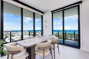A dining area with a large table and chairs, large windows with a city and ocean view, and a balcony with a chair.