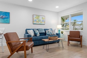 Ein Wohnzimmer mit einem blauen Sofa, zwei braunen Sesseln, einem Holztisch und einem Fenster mit Blick auf Palmen.