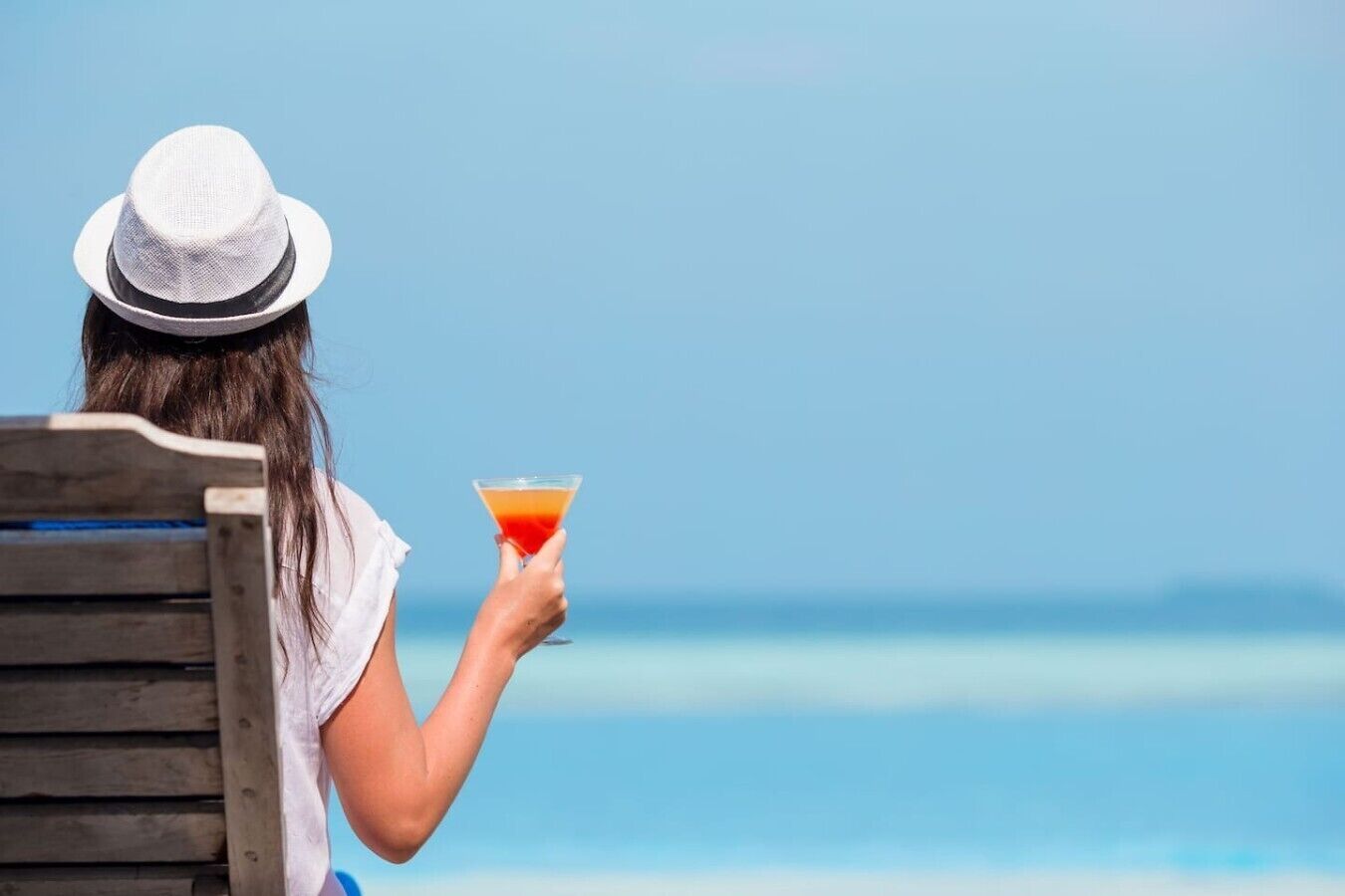 Eine Person sitzt mit einem Getränk in der Hand auf einer Holzbank und blickt auf das ruhige Meer.