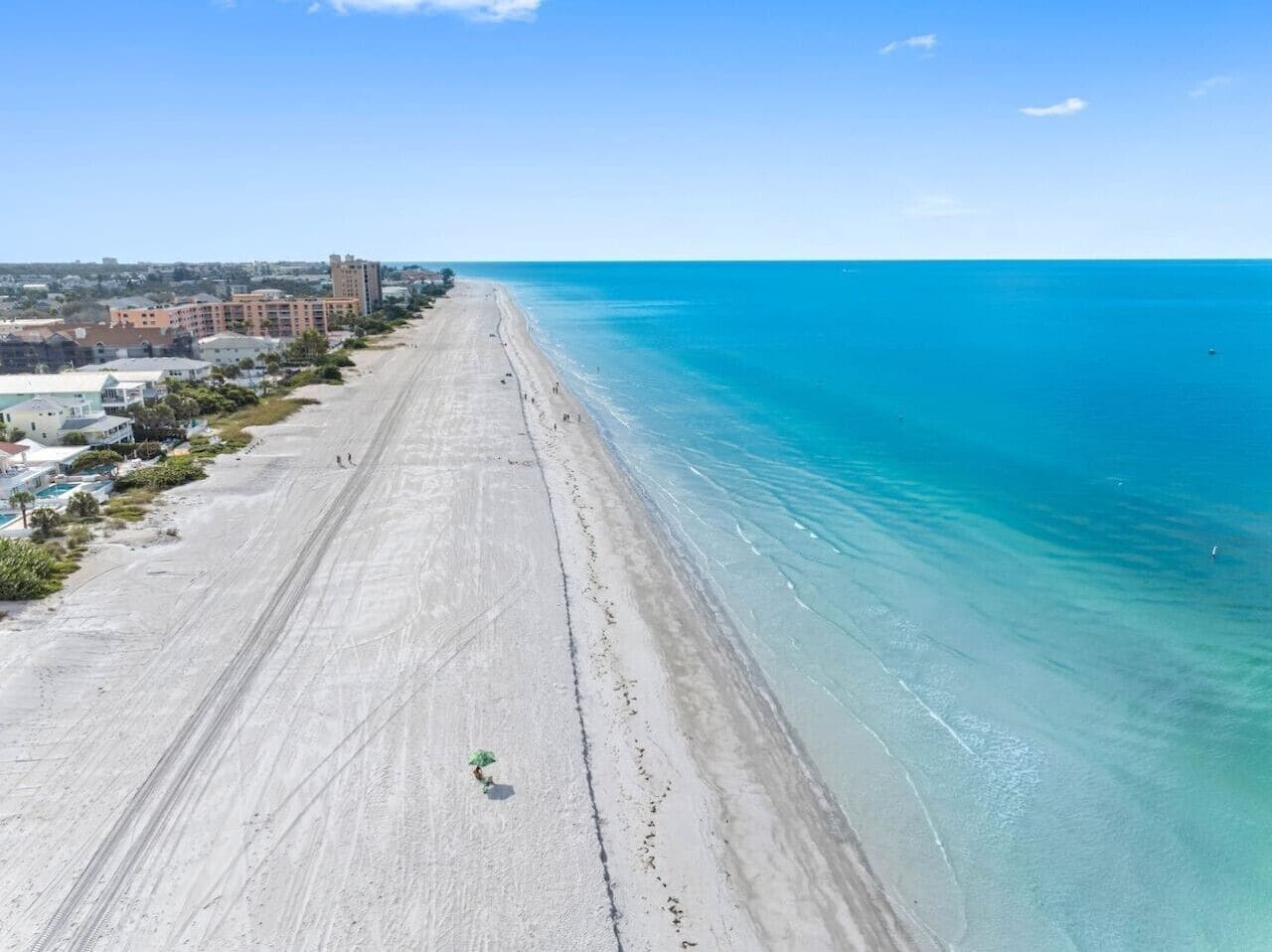 A long, wide beach with clear blue water and a few buildings in the background.