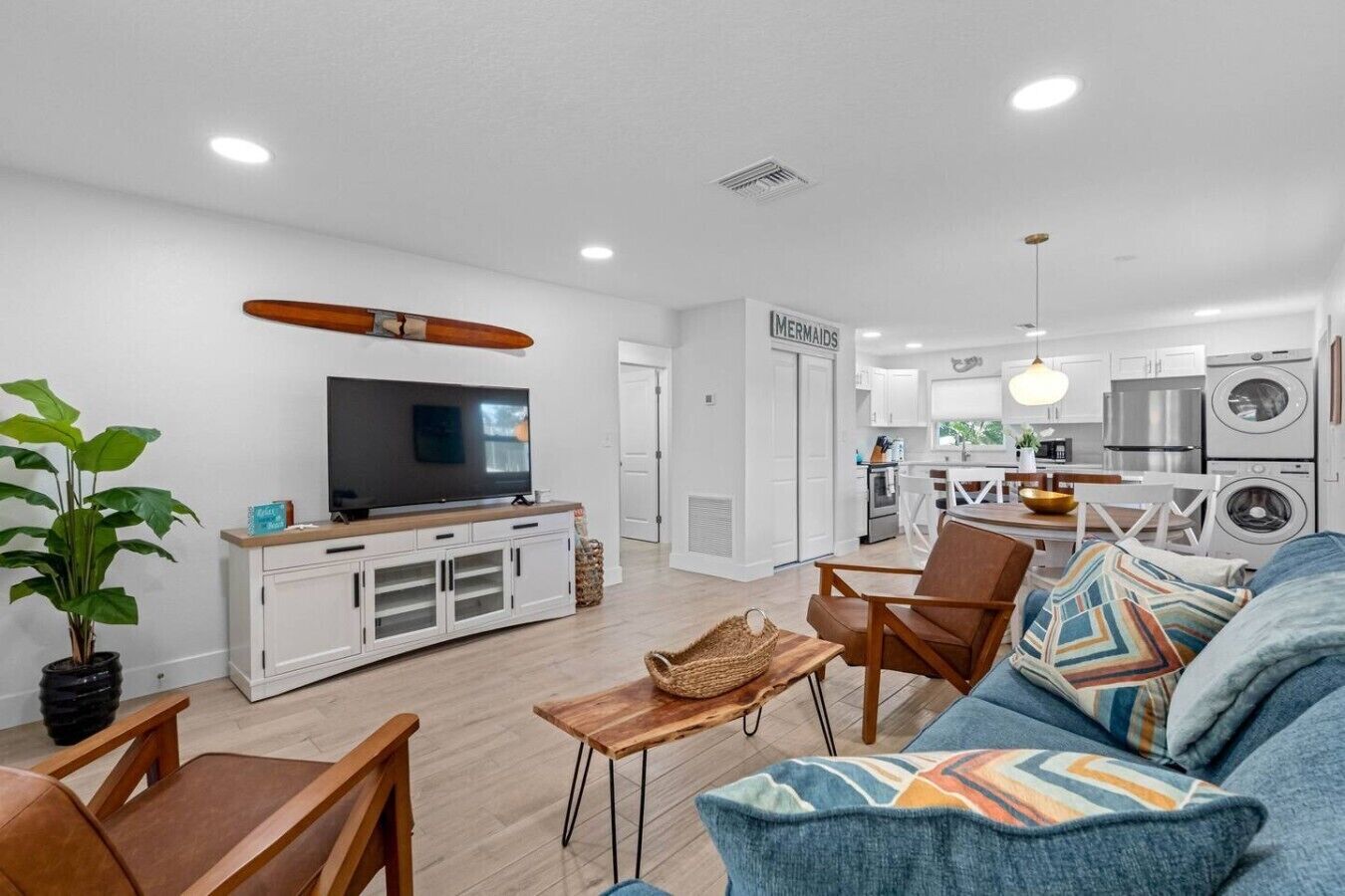 A modern living room with a flat-screen TV, wooden furniture, a sofa with patterned cushions, and a dining area with a table and chairs.