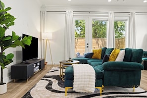 Een moderne woonkamer met een hoekbank, een flatscreen televisie, een vloerlamp en een groot raam met gordijnen.