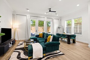 A modern living room with a sectional sofa, a flat-screen TV, a dining area with a table and chairs, and large windows with a view of greenery. - The Suite Spot: 4BR with 2 King Suites and Private Patio near NRG Stadium (Houston)