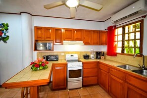 A kitchen with wooden cabinets, a microwave, a stove, a sink, and a window with red curtains. - Garden Paradise, Walk to the Beach (Tamarindo)