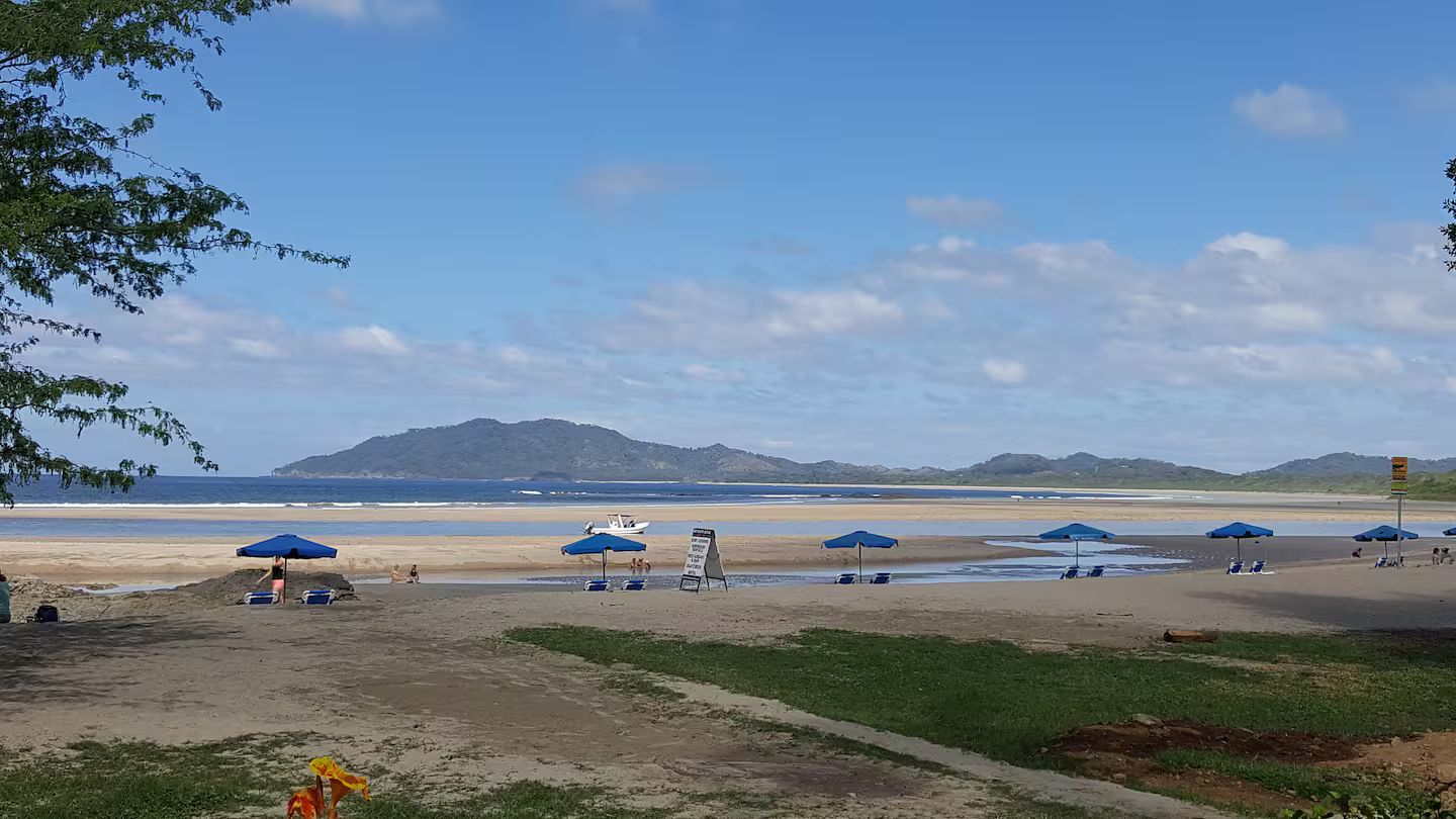 Una playa con sombrillas y un bote, cielo despejado y montañas a lo lejos.