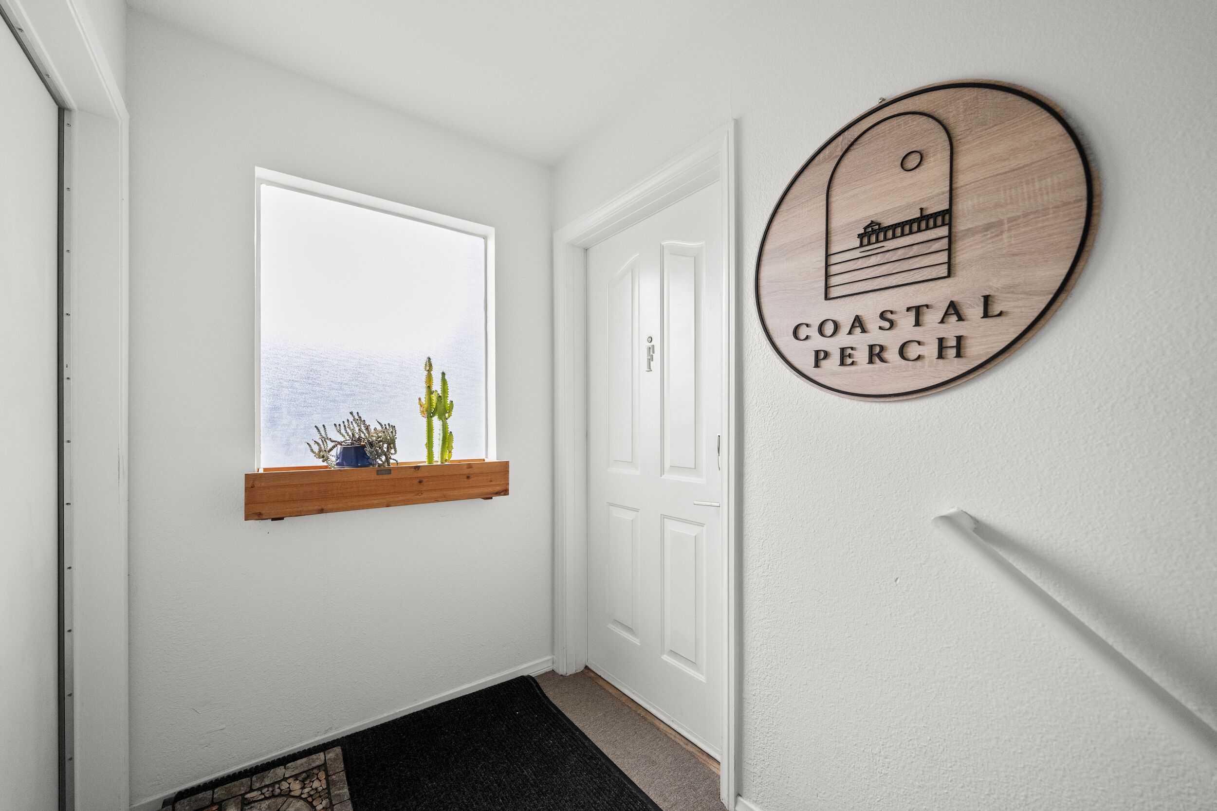 Ein Flur mit einer weißen Tür, einem Fenster mit Holzregal und einem an der Wand befestigten Schild mit der Aufschrift 'COASTAL PERCH'.