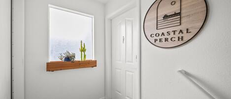 Um corredor com uma porta branca, uma janela com prateleira de madeira e uma placa na parede com a inscrição 'COASTAL PERCH'.