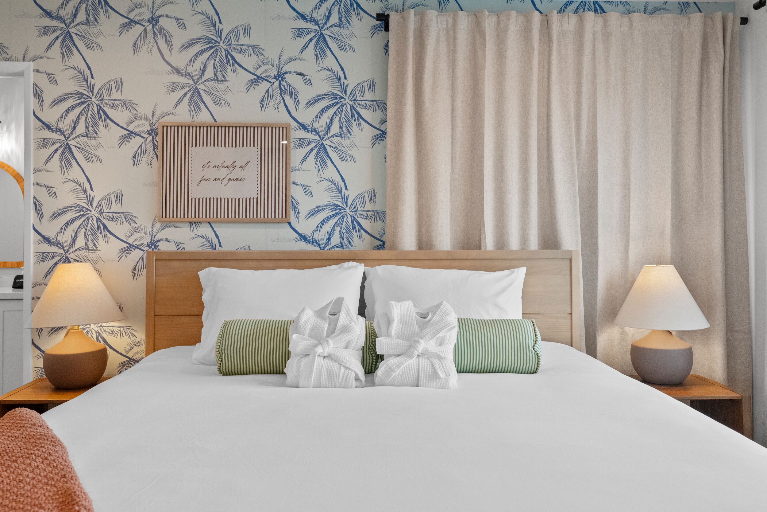 A hotel room with a bed, pillows, a headboard, a nightstand, a lamp, and a wall with a palm tree pattern.