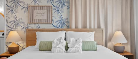 A hotel room with a bed, pillows, a headboard, a nightstand, a lamp, and a wall with a palm tree pattern.